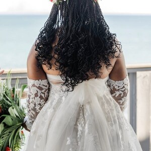 May include: A bride wearing a white lace wedding dress with a floral crown. The bride is standing with her back to the camera, looking out at the ocean.