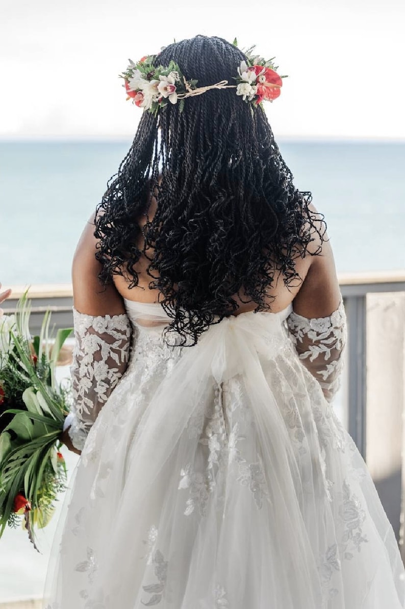 May include: A bride wearing a white lace wedding dress with a floral crown. The bride is standing with her back to the camera, looking out at the ocean.