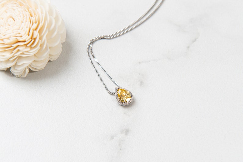 Pu&ograve; includere: Una collana a catena d'argento con un ciondolo a forma di pera in pietra preziosa gialla circondato da piccole pietre preziose trasparenti.