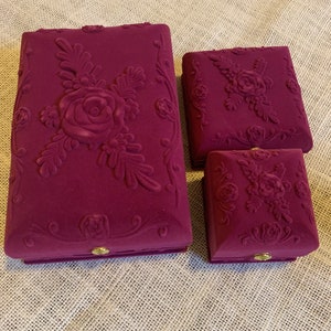 Pu&ograve; includere: Tre scatole per gioielli in velluto con motivi floreali. La scatola pi&ugrave; grande &egrave; color borgogna con un grande disegno di rosa e foglie. Le due scatole pi&ugrave; piccole sono quadrate e hanno un disegno di rosa e foglie pi&ugrave; piccolo.