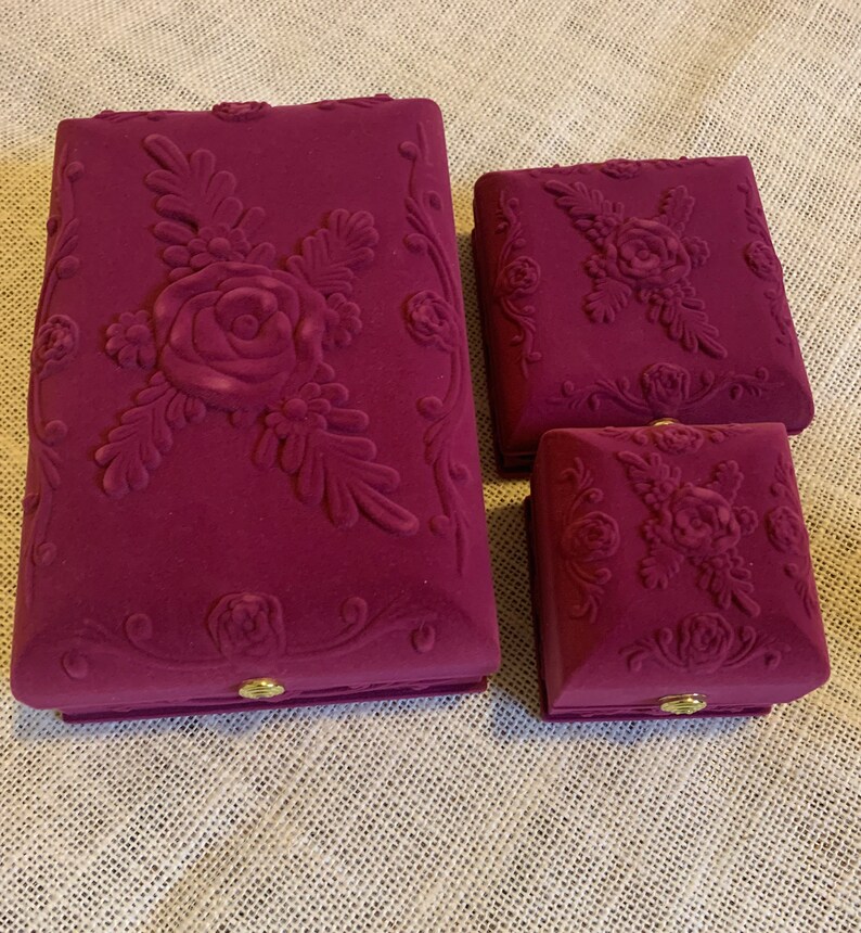 Pu&ograve; includere: Tre scatole per gioielli in velluto con motivi floreali. La scatola pi&ugrave; grande &egrave; color borgogna con un grande disegno di rosa e foglie. Le due scatole pi&ugrave; piccole sono quadrate e hanno un disegno di rosa e foglie pi&ugrave; piccolo.