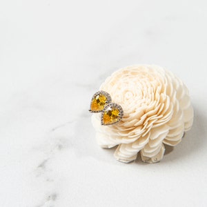 Pu&ograve; includere: Un paio di orecchini gialli a forma di pera con un alone di pietre chiare, posizionati su un fiore bianco.
