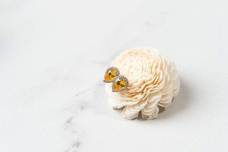 Pu&ograve; includere: Un paio di orecchini gialli a forma di pera con un alone di pietre chiare, posizionati su un fiore bianco.