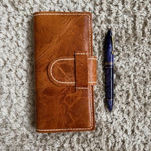 May include: A brown leather wallet with a white stitch detail and a flap closure. A blue pen is lying next to the wallet on a light brown fuzzy surface.