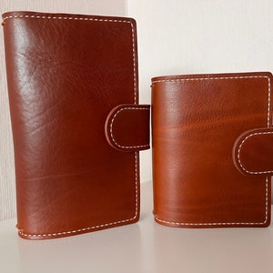 May include: Two brown leather bound journals with white stitching. The larger journal has a flap closure. Both journals are on a white surface.