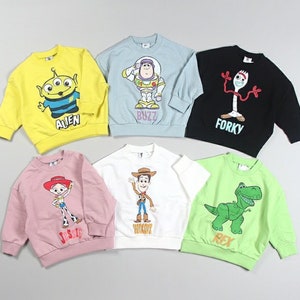 Puede incluir: Seis sudaderas de colores con personajes de la franquicia Toy Story. Las sudaderas son amarillas, azul claro, negras, rosas, blancas y verde claro. Cada sudadera presenta un personaje diferente, incluyendo Buzz Lightyear, Woody, Jessie, Rex, Forky y el Alien.