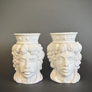 Può includere: Due vasi bianchi in ceramica a forma di volto. Un vaso ha i baffi e l'altro no. Entrambi i vasi hanno una corona e dettagliati tratti del viso. I vasi sono su una superficie grigio scuro.