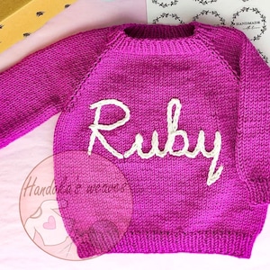 K&ouml;nnte beinhalten: Ein leuchtend magentafarbener, lang&auml;rmeliger Strickpullover mit dem Namen Ruby, der in cremefarbenem Garn auf die Brust gestickt ist. Das Kleidungsst&uuml;ck hat einen gerippten Rundhalsausschnitt, gerippte B&uuml;ndchen und einen gerippten Saum.