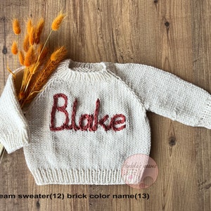 K&ouml;nnte beinhalten: Ein cremefarbener, lang&auml;rmeliger Strickpullover mit dem eingestickten Namen Blake in ziegelrotem Garn auf der Brust. Der Pullover hat einen gerippten Rundhalsausschnitt, B&uuml;ndchen und Saum und ist mit einem kleinen B&uuml;ndel getrocknetem orangefarbenem Gras dekoriert.