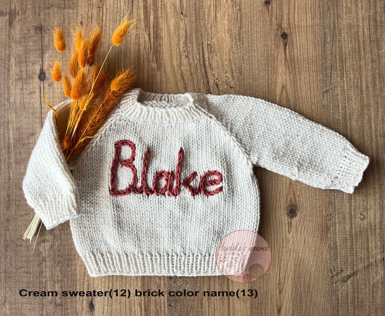 K&ouml;nnte beinhalten: Ein cremefarbener, lang&auml;rmeliger Strickpullover mit dem eingestickten Namen Blake in ziegelrotem Garn auf der Brust. Der Pullover hat einen gerippten Rundhalsausschnitt, B&uuml;ndchen und Saum und ist mit einem kleinen B&uuml;ndel getrocknetem orangefarbenem Gras dekoriert.