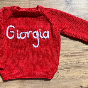 K&ouml;nnte beinhalten: Ein leuchtend roter, lang&auml;rmeliger Strickpullover mit dem Namen Giorgia, der in wei&szlig;er Schreibschrift auf die Brust gestickt ist. Das Kleidungsst&uuml;ck hat einen gerippten Rundhalsausschnitt, gerippte B&uuml;ndchen und einen gerippten Saum.