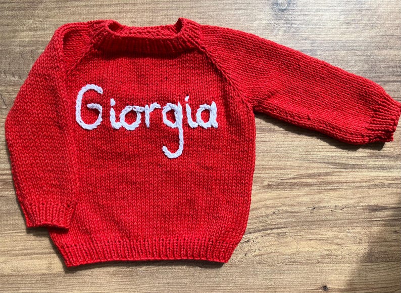 K&ouml;nnte beinhalten: Ein leuchtend roter, lang&auml;rmeliger Strickpullover mit dem Namen Giorgia, der in wei&szlig;er Schreibschrift auf die Brust gestickt ist. Das Kleidungsst&uuml;ck hat einen gerippten Rundhalsausschnitt, gerippte B&uuml;ndchen und einen gerippten Saum.