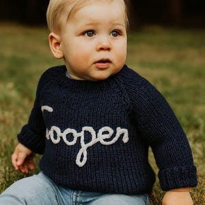 K&ouml;nnte beinhalten: Ein dunkelblauer, lang&auml;rmeliger Strickpullover mit dem Namen Cooper in wei&szlig;er Schreibschrift auf der Brust, kombiniert mit einer hellblauen Jeans. Der Pullover hat eine gerippte Struktur und umgeschlagene &Auml;rmelb&uuml;ndchen.
