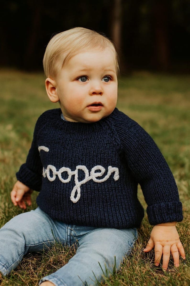 K&ouml;nnte beinhalten: Ein dunkelblauer, lang&auml;rmeliger Strickpullover mit dem Namen Cooper in wei&szlig;er Schreibschrift auf der Brust, kombiniert mit einer hellblauen Jeans. Der Pullover hat eine gerippte Struktur und umgeschlagene &Auml;rmelb&uuml;ndchen.