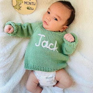 K&ouml;nnte beinhalten: Ein lang&auml;rmeliger, salbeigr&uuml;ner Strickpullover mit dem in wei&szlig;em Garn auf die Brust gestickten Namen Jack. Der Pullover hat einen gerippten Halsausschnitt, B&uuml;ndchen und Saum. Eine runde Meilensteinscheibe aus Holz mit Dinosaurier-Motiv und Text 1 MONTH.