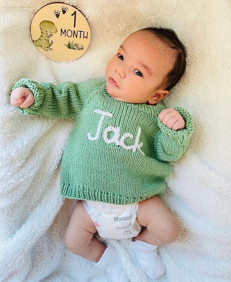 K&ouml;nnte beinhalten: Ein lang&auml;rmeliger, salbeigr&uuml;ner Strickpullover mit dem in wei&szlig;em Garn auf die Brust gestickten Namen Jack. Der Pullover hat einen gerippten Halsausschnitt, B&uuml;ndchen und Saum. Eine runde Meilensteinscheibe aus Holz mit Dinosaurier-Motiv und Text 1 MONTH.