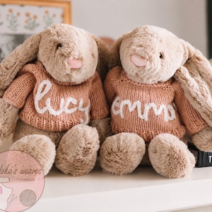 May include: Two plush beige rabbits with long floppy ears and pink noses, each wearing a peach-colored knitted sweater. The left sweater features the white embroidered name "lucy" and the right sweater features the white embroidered name "emma".