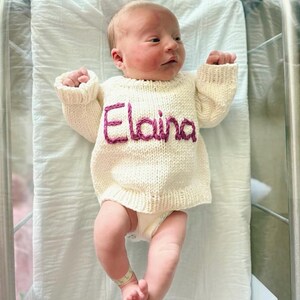 K&ouml;nnte beinhalten: Ein cremefarbener, lang&auml;rmeliger Strickpullover mit dem Namen Elaina, der in dunkelrosa Garn auf die Brust gestickt ist. Der Pullover hat einen gerippten Halsausschnitt, B&uuml;ndchen und Saum und wird von einem Neugeborenen auf einem wei&szlig;en Krankenhauslaken getragen.
