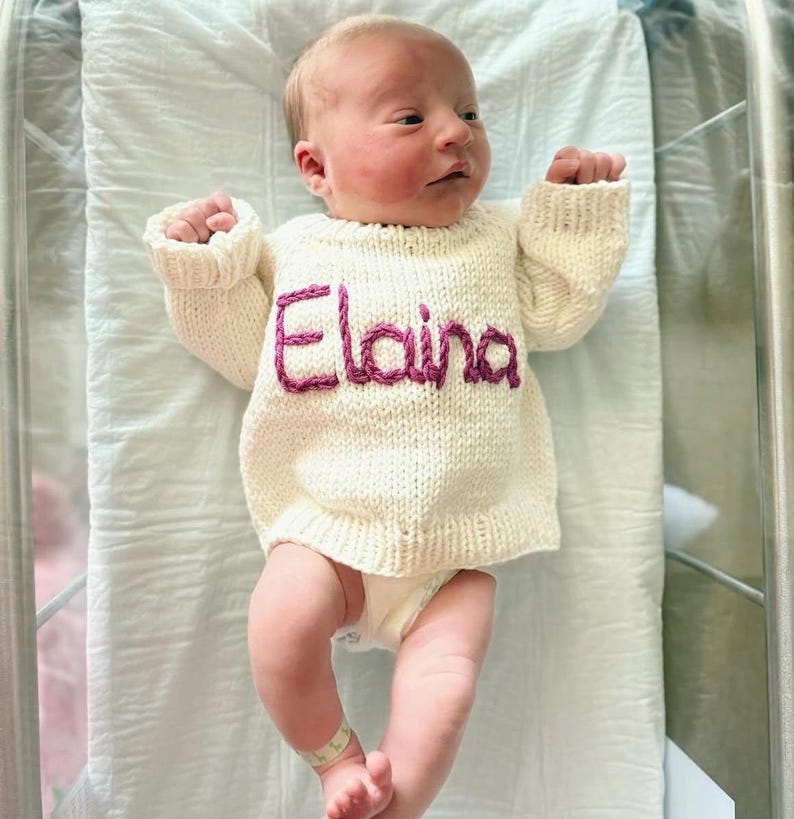 K&ouml;nnte beinhalten: Ein cremefarbener, lang&auml;rmeliger Strickpullover mit dem Namen Elaina, der in dunkelrosa Garn auf die Brust gestickt ist. Der Pullover hat einen gerippten Halsausschnitt, B&uuml;ndchen und Saum und wird von einem Neugeborenen auf einem wei&szlig;en Krankenhauslaken getragen.