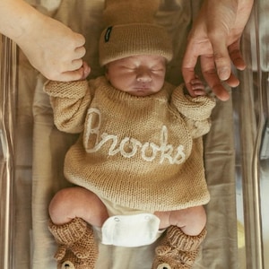 K&ouml;nnte beinhalten: Ein Neugeborenes tr&auml;gt einen beigen Strickpullover mit dem in wei&szlig;er Schreibschrift aufgestickten Namen Brooks, eine passende beige Strickm&uuml;tze und beige Stricksch&uuml;hchen mit einem B&auml;rengesicht-Motiv, kombiniert mit einer wei&szlig;en Windel.