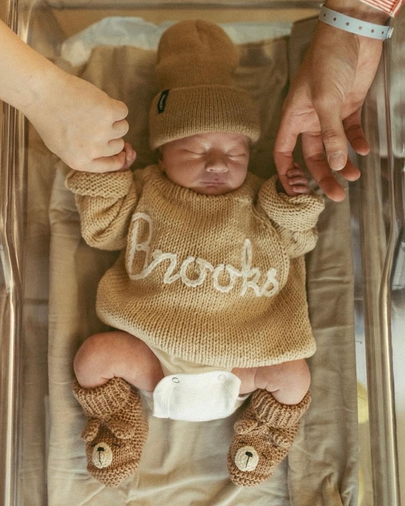 K&ouml;nnte beinhalten: Ein Neugeborenes tr&auml;gt einen beigen Strickpullover mit dem in wei&szlig;er Schreibschrift aufgestickten Namen Brooks, eine passende beige Strickm&uuml;tze und beige Stricksch&uuml;hchen mit einem B&auml;rengesicht-Motiv, kombiniert mit einer wei&szlig;en Windel.