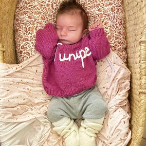 K&ouml;nnte beinhalten: Ein Baby tr&auml;gt einen magentafarbenen, lang&auml;rmeligen Strickpullover mit dem wei&szlig;en, gestickten Schriftzug "Juniper" auf der Brust, dazu eine graue Hose und cremefarbene weiche Babyschuhe. Das Baby liegt auf einem gemusterten Stoff in einem Korb.