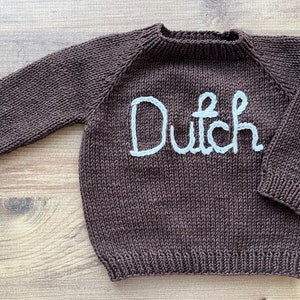 K&ouml;nnte beinhalten: Ein dunkelbrauner, lang&auml;rmeliger Strickpullover mit dem Wort Dutch, das in wei&szlig;em Garn auf die Brust gestickt ist. Das Kleidungsst&uuml;ck verf&uuml;gt &uuml;ber einen gerippten Rundhalsausschnitt, gerippte B&uuml;ndchen und einen gerippten Saum im Glattstrick.