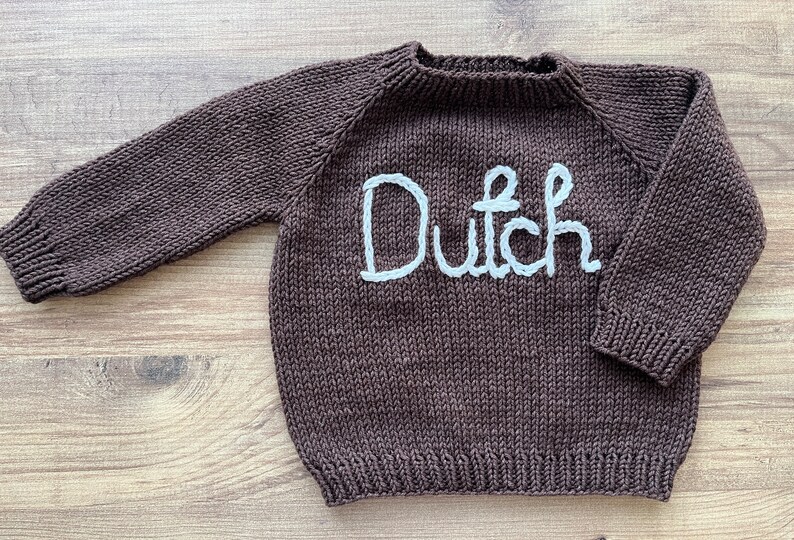 K&ouml;nnte beinhalten: Ein dunkelbrauner, lang&auml;rmeliger Strickpullover mit dem Wort Dutch, das in wei&szlig;em Garn auf die Brust gestickt ist. Das Kleidungsst&uuml;ck verf&uuml;gt &uuml;ber einen gerippten Rundhalsausschnitt, gerippte B&uuml;ndchen und einen gerippten Saum im Glattstrick.