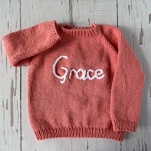 K&ouml;nnte beinhalten: Ein korallenfarbener, lang&auml;rmeliger Strickpullover mit Rundhalsausschnitt sowie gerippten B&uuml;ndchen an &Auml;rmeln und Saum. Auf der Vorderseite ist der Name Grace in wei&szlig;er Schreibschrift mittig auf der Brust eingestickt.