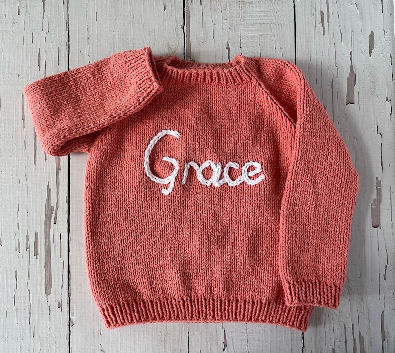 K&ouml;nnte beinhalten: Ein korallenfarbener, lang&auml;rmeliger Strickpullover mit Rundhalsausschnitt sowie gerippten B&uuml;ndchen an &Auml;rmeln und Saum. Auf der Vorderseite ist der Name Grace in wei&szlig;er Schreibschrift mittig auf der Brust eingestickt.
