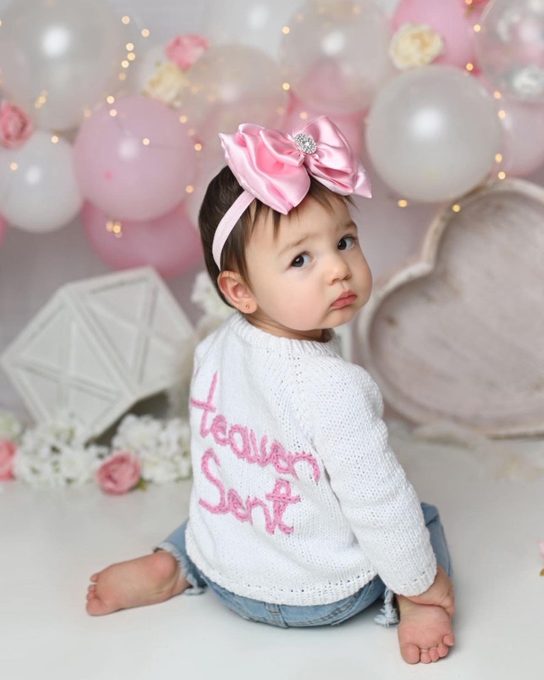 K&ouml;nnte beinhalten: Ein wei&szlig;er, lang&auml;rmeliger Strickpullover mit dem Schriftzug "Heaven Sent", der in rosa Garn auf den R&uuml;cken gestickt ist, kombiniert mit einer blauen Jeans. Eine gro&szlig;e rosa Haarschleife aus Satin mit einem zentralen Strassstein an einem d&uuml;nnen rosa Haarband.