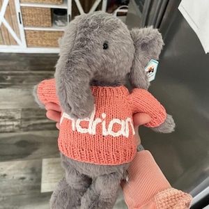 May include: A grey plush elephant toy wearing a coral-coloured knitted jumper. The jumper has the name "Adrian" in white letters. The elephant has large ears and a tag attached.