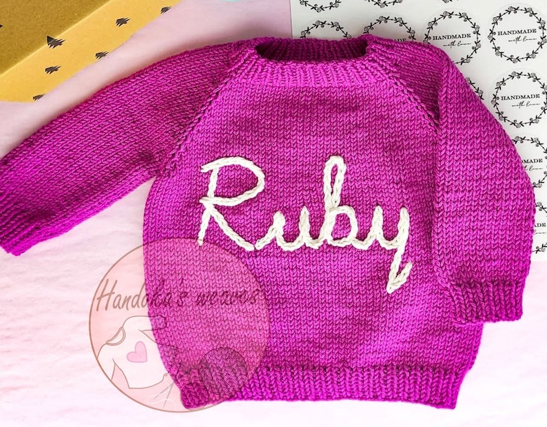 K&ouml;nnte beinhalten: Ein leuchtend magentafarbener, lang&auml;rmeliger Strickpullover mit dem Namen Ruby, der in cremefarbenem Garn auf die Brust gestickt ist. Das Kleidungsst&uuml;ck hat einen gerippten Rundhalsausschnitt, gerippte B&uuml;ndchen und einen gerippten Saum.