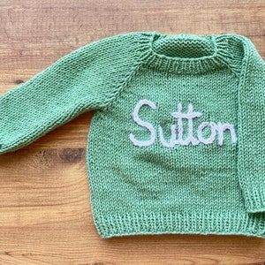 K&ouml;nnte beinhalten: Ein salbeigr&uuml;ner, lang&auml;rmeliger Strickpullover mit Rundhalsausschnitt und gerippten B&uuml;ndchen. Auf der Vorderseite des Kleidungsst&uuml;cks ist der Name Sutton in wei&szlig;em Garn in Schreibschrift aufgestickt, mittig auf dem Brustbereich des Strickstoffs.