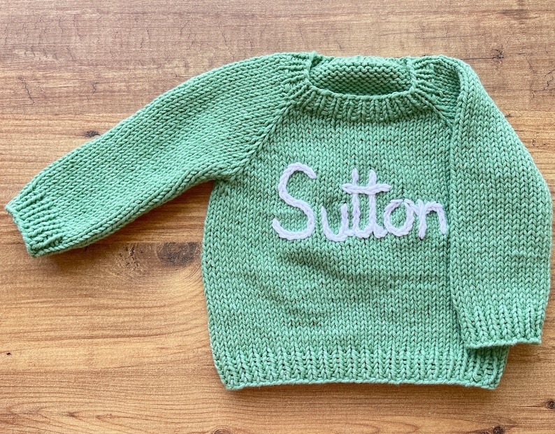 K&ouml;nnte beinhalten: Ein salbeigr&uuml;ner, lang&auml;rmeliger Strickpullover mit Rundhalsausschnitt und gerippten B&uuml;ndchen. Auf der Vorderseite des Kleidungsst&uuml;cks ist der Name Sutton in wei&szlig;em Garn in Schreibschrift aufgestickt, mittig auf dem Brustbereich des Strickstoffs.