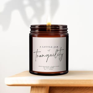 Peut inclure: Une bougie en cire de soja allumée dans un pot en verre brun. L'étiquette indique "A Little Jar of Tranquility" avec "Rose Geranium + Ylang Ylang" et "6oz / 180ml". La bougie est posée sur une surface en bois.