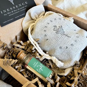 A gift box containing a small glass bottle with a label, a white cloth bag with a floral design, and some shredded paper.