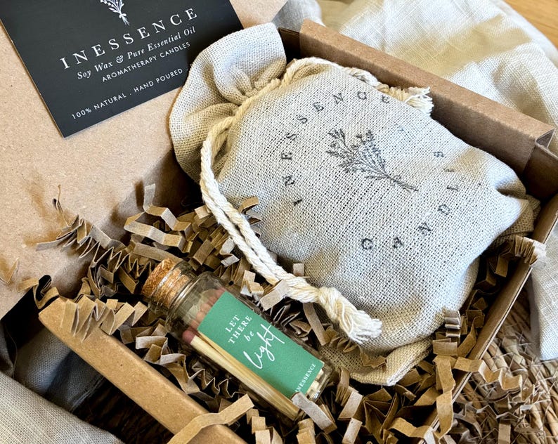 A gift box containing a small glass bottle with a label, a white cloth bag with a floral design, and some shredded paper.