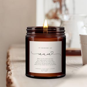 Puede incluir: Una vela de cera de soja encendida en un tarro de cristal marrón. La vela tiene una etiqueta blanca con el texto "IN MEMORY OF nan" y un poema. Perfumada con manzanilla, ylang ylang y lavanda. El tarro es de 170 ml.