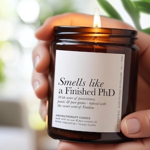 May include: A lit aromatherapy candle in a brown glass jar, held in a hand. The candle has a white label with the text "Smells like a Finished PhD". The label also lists the scent notes and ingredients. The candle is 180ml.