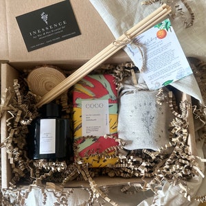 Op de afbeelding: Een cadeaudoos gevuld met bruin papierstrooisel, een zwarte glazen fles met een wit etiket, een set houten diffuserstokjes, een kleine ronde houten schijf, een reep chocolade verpakt in kleurrijk papier en een kleine linnen tas met een logo.