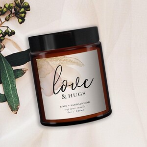 Op de afbeelding: Een bruine glazen potkaars met een zwart deksel. Het etiket luidt "love & HUGS" met "ROSE + SANDALWOOD" en "6oz / 180ml" eronder. De achtergrond heeft een zacht, neutraal kleurenpalet met bloemaccenten.