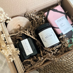Könnte beinhalten: Eine Geschenkbox gefüllt mit braunem Packpapier und einem braunen und weißen Holz-Untersetzer. Die Box enthält zwei schwarze Glaskerzen, eine mit dem Text "Mum" und die andere mit dem Text "wild & fragrant". Außerdem gibt es eine rosa und schwarze Schokoladentafel mit dem Text "Coco" und den Worten "Lavender Milk Chocolate".