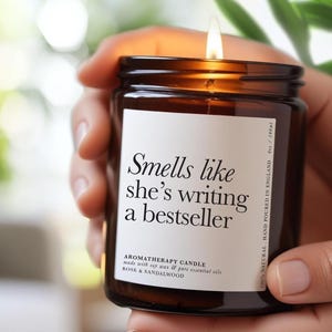 Peut inclure: Une bougie d'aromathérapie allumée dans un pot en verre brun. L'étiquette indique "Smells like she's writing a bestseller". La bougie est faite de cire de soja et d'huiles essentielles de rose et de bois de santal. Le pot est de 180 ml.