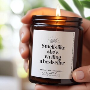 May include: A lit aromatherapy candle in a brown glass jar with a white label featuring the text Smells like she's writing a bestseller, Aromatherapy Candle, made with love, soy wax & pure essential oils, 100% natural, hand poured in England, 6oz / 180ml.
