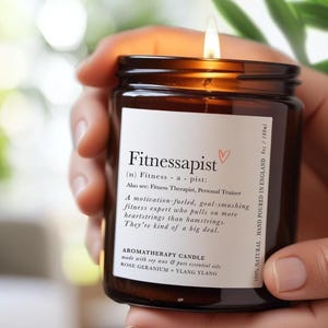 May include: A lit aromatherapy candle in a brown glass jar, held by a person. The label reads "Fitnessapist" with a definition and details about the candle's soy wax, essential oils, and origin. The candle is 180ml.