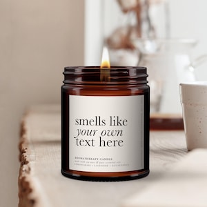 Peut inclure: Une bougie d'aromathérapie dans un pot en verre brun avec une mèche allumée et une étiquette blanche portant le texte noir : smells like your own text here, aromatherapy candle, made with soy wax & pure essential oils, lemongrass + lavender + eucalyptus.