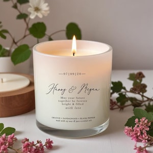 May include: A burning white candle in a clear glass jar. The candle is inscribed with "Harry & Megan" and a message of love. The candle's scent is orange, sandalwood, and black pepper. The candle is made with soy wax and pure essential oils.