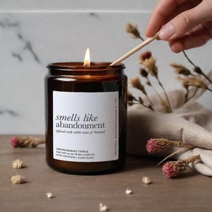 May include: A lit aromatherapy candle in a brown glass jar with a white label reading "smells like abandonment" and "infused with subtle notes of betrayal." The candle is made with soy wax and essential oils of rose geranium and ylang ylang.