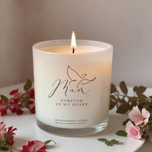May include: A lit candle in a clear glass jar with a white label featuring a black line drawing of a bird above the text Mum, Forever in My Heart, and Aromatherapy Candle, made with soy wax and pure essential oils, glowing with a warm flame.